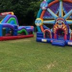 Two colorful inflatable structures—a carnival-themed bounce house Long Island and an obstacle course—are set up on grass outdoors with signs showing entry prices. Trees and a fence are visible in the background, perfect for party rentals Long Island, NY.