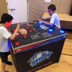 Two boys play a reaction game, hitting illuminated buttons on a large arcade-style machine at a party rentals Long Island event in a gymnasium. The game has red and blue lights and a digital score display.