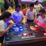 Four children in school uniforms play a large electronic reaction game with colored buttons in a gymnasium, while other kids and party rentals Long Island enjoy nearby inflatables in the background.