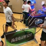Children in gym uniforms play electronic games in a gym; one girl tries the “Megawire” game, while others enjoy a light-up arcade—perfect inspiration for bounce house Long Island parties or fun party rentals Long Island, NY.