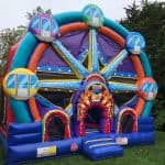 A colorful inflatable bounce house Long Island shaped like a Ferris wheel with cartoon gondolas, a clown face entrance, and festive decorations, set up on green grass with trees in the background—perfect for party rentals Long Island, NY.