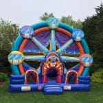 A colorful inflatable bounce house Long Island shaped like a Ferris wheel sits on a grassy lawn, surrounded by trees and bushes, with a cloudy sky overhead—perfect for NY party rentals.