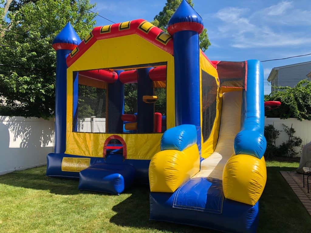 A colorful inflatable bounce house with a slide sits on green grass in a backyard, surrounded by trees and a white fence under a blue sky—perfect for party rentals Long Island celebrations.