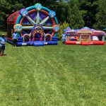 A grassy field features carnival games, inflatable attractions like a bounce house Long Island partygoers love, and two red-and-white tents. Four staff in blue shirts welcome guests, with trees and outdoor seating completing this NY event scene.
