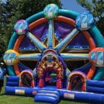 A man stands beside a large, colorful bounce house Long Island designed to look like a Ferris wheel, set up on a grassy lawn with trees in the background.