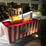 A popcorn machine sits on a red-and-white striped table outdoors at night. Several tubs of popcorn are lined up under a sign reading "The Big Popcorn," perfect for party rentals Long Island, NY. Plants can be seen in the background.