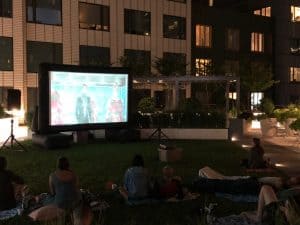People sit on blankets on a grassy lawn at night, watching a movie projected on an outdoor screen. Apartment buildings and plants are visible in the background, bringing a relaxed, social vibe—perfect for party rentals Long Island gatherings.