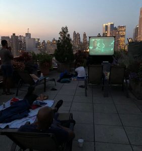 People watch a movie on a rooftop terrace at dusk, lounging in chairs with blankets. A projector screen plays the film against a city skyline—perfect inspiration for unique party rentals Long Island, NY has to offer.