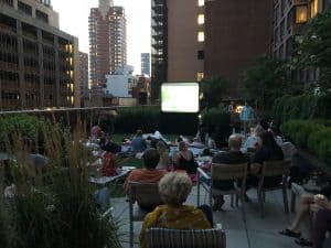 People sit on chairs and blankets on a rooftop garden at dusk, watching a movie projected on a screen amid city buildings and greenery—a perfect scene for party rentals Long Island, NY residents might choose for unique gatherings.
