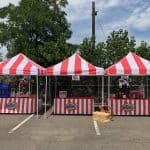 Three red-and-white striped carnival tents set up in a parking lot, each with a counter and fun decorations—perfect for party rentals Long Island. Hay bales and red wagon wheels add charm, with trees and a fence in the background.
