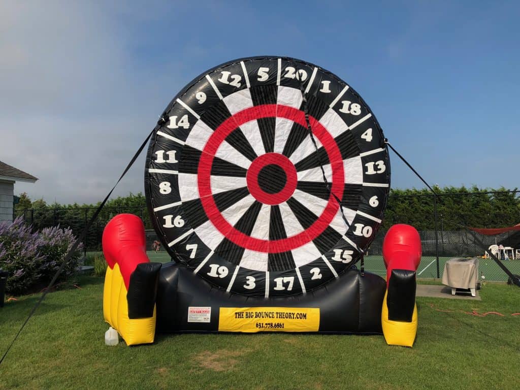 A giant inflatable dartboard stands outdoors on grass, perfect for party rentals Long Island. With red, black, yellow, and white sections, it’s supported by black cords and flanked by two red and yellow inflatable gloves at the base.