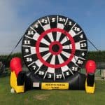 A giant inflatable dartboard stands outdoors on grass, perfect for party rentals Long Island. With red, black, yellow, and white sections, it’s supported by black cords and flanked by two red and yellow inflatable gloves at the base.