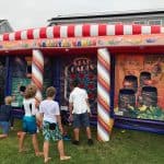 Several children play games at an outdoor inflatable carnival booth with colorful decorations and banners. Nearby, a bounce house Long Island adds extra fun for the kids, making this event a highlight for party rentals Long Island, NY!.