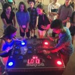 A group of teens gathers around a large, illuminated arcade game table at a party rentals Long Island event. Two players, one in a tank top and one in glasses, compete by pressing glowing buttons as the others watch and smile.
