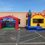 Two inflatable play structures are set up in a parking lot: a red and blue Game Zone tent and a colorful castle-shaped bounce house. Perfect for party rentals Long Island, the setup is in front of a beige building.