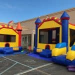 Two colorful inflatable bounce houses with turrets and slides are set up outside on a paved area near a building under the blue sky—perfect for any party rentals Long Island event.