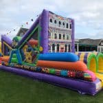 A colorful inflatable obstacle course with arches and patterns sits on a grassy lawn—perfect for party rentals Long Island—with houses and outdoor tables in the background under a cloudy sky.