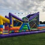 A colorful inflatable obstacle course, perfect for party rentals Long Island, sits on green grass outdoors with people nearby and bunting flags above. The bright purple, yellow, blue, and red sections surround a sign that reads "COME ONE COME ALL.