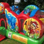A colorful inflatable obstacle course with cartoon animals, including monkeys, elephants, and giraffes. An adult helps a young child enter the bounce house Long Island on a grassy lawn during a festive NY party.