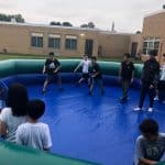 A group of kids play a game inside an inflatable bounce house Long Island arena outdoors, with school buildings in the background. Some are barefoot and actively engaged, while others stand on the sidelines watching.