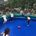 Children play Gaga ball inside an inflatable octagon outdoors, while a crowd of kids and adults watch from around the enclosure. Trees and tents are visible in the background at a community event with fun party rentals Long Island, NY.