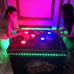 Two women, one in a wedding dress and the other in black, play an illuminated "Strike a Light" arcade game in a dimly lit room—perfect inspiration for party rentals Long Island, NY events or adding fun beyond just a bounce house Long Island.