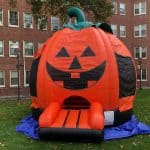 An inflatable orange pumpkin bounce house Long Island with a jack-o'-lantern face serves as an entrance on a grassy lawn, with a brick building and trees in the background.