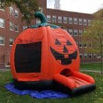 A large inflatable pumpkin bounce house Long Island with a jack-o’-lantern face stands on a blue tarp in front of a brick school building and trees on a grassy lawn, perfect for party rentals Long Island, NY.