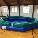 A large, inflatable octagonal arena with green and blue sides sits on a gymnasium floor under high windows and wooden beams—ideal for party rentals Long Island, NY or as a unique bounce house Long Island event centerpiece.