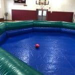 An empty indoor gym features a large, inflatable, octagonal arena—like those found in bounce house Long Island, NY party rentals—with blue flooring and green walls; a single red ball sits in the center. A basketball hoop and scoreboard are visible in the background.