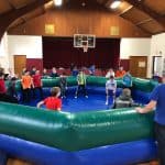 Children and adults gather inside a large inflatable gaga ball pit—popular among party rentals Long Island—in a gymnasium with a basketball hoop, wooden ceiling, and stage. Several kids are ready to play while staff supervise.