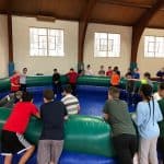 A group of kids and teens stand around and inside a large, inflatable octagonal arena in a gymnasium—possibly from party rentals Long Island—as sunlight streams in through the windows above, ready to play a game.