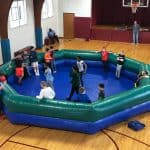 Children and adults gather around and inside a large inflatable octagonal arena—perfect for party rentals Long Island—in a gymnasium, possibly preparing to play a group game. A basketball hoop and bleachers are visible in the background.