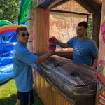 Two people in blue shirts stand at a tiki-themed inflatable booth outdoors from a party rentals Long Island company; one hands the other a red cup. Inflatable palm trees and a rainbow are visible in the background on a sunny day.