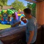 Two people in blue shirts stand by a bounce house Long Island structure; one pours a drink from a red cooler into the other's cup. Colorful inflatable slides and greenery complete this NY outdoor party rentals Long Island scene.
