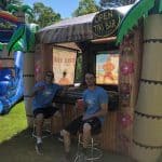Two men sit on stools in front of an inflatable tiki bar with an "OPEN TIKI BAR" sign, holding red cups. A large bounce house Long Island water slide is visible in the background on a sunny day.