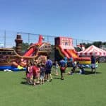 Children and adults gather on a grassy field near bounce house Long Island, NY inflatables shaped like a pirate ship and circus tent, with a clear blue sky and sports fence in the background.