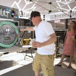 A man in a white shirt and cap plays "The Vault," an arcade-style game from party rentals Long Island, outdoors while two women stand nearby smiling and holding drinks. The scene feels casual and festive under a canopy, perfect for NY gatherings.