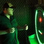 A man wearing a cap and plaid shirt stands in front of a circular electronic game with glowing green lights, holding a handheld controller and looking focused—perfect inspiration for party rentals Long Island celebrations.