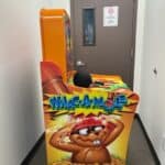 A Whac-A-Mole arcade game in a narrow hallway, ideal for NY party rentals Long Island, sits beside a closed brown door with a "No Smoking or Vaping" sign. The game's side art features a cartoon mole wearing a red helmet.