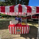 Two men stand under a red and white striped canopy at an outdoor event in NY, behind a table with cotton candy and popcorn machines. Trees and bounce house Long Island inflatables are in the background at this festive scene.