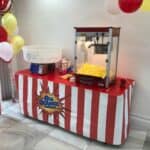 A red-and-white striped table holds a popcorn machine full of popcorn, a cotton candy maker, bags of popcorn, and yellow and red balloons—perfect for an NY celebration or pairing with bounce house Long Island party rentals.