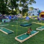 A mini-golf course set up on grass with several people playing and socializing nearby. In the background, a colorful bounce house Long Island attraction, trees, and large white buildings stand under a clear blue sky.