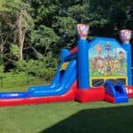 A brightly colored inflatable bounce house and slide with a Paw Patrol theme sits on green grass in a sunny backyard, perfect for party rentals Long Island celebrations, with trees and a wooden playset in the background.