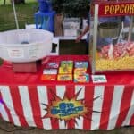 A concession stand with a red and white striped tablecloth offers popcorn, cotton candy, and boxed candies like M&M’s and Skittles. The sign reads “The Big Bounce Theory”—perfect for bounce house Long Island party rentals in NY.