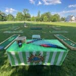 A green striped table with mini golf clubs, balls, and scorecards sits on a grassy field in NY, with several mini golf holes set up in the background under a blue sky—perfect for party rentals Long Island gatherings.
