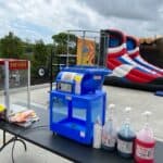 A table with a popcorn machine, a blue cotton candy machine, and syrup bottles sits outdoors near bounce house Long Island inflatables shaped like basketball shoes and a log cabin. An American flag waves to the left under a cloudy sky.