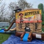 An inflatable western-themed bounce house Long Island with a slide, decorated with cowboy, cactus, and saloon imagery, set up on grass with a blue tarp—perfect for backyard fun and party rentals Long Island, NY.