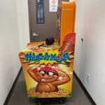 An orange Whac-A-Mole arcade machine sits in a narrow hallway, perfect for party rentals Long Island events. A cartoon mole wearing a red headband decorates its side, while a closed brown door with a no smoking sign is in the background.