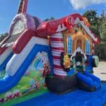A colorful inflatable bounce house Long Island shaped like a gingerbread house with candy decorations and a slide stands on a blue tarp outdoors, surrounded by trees under a partly cloudy NY sky.